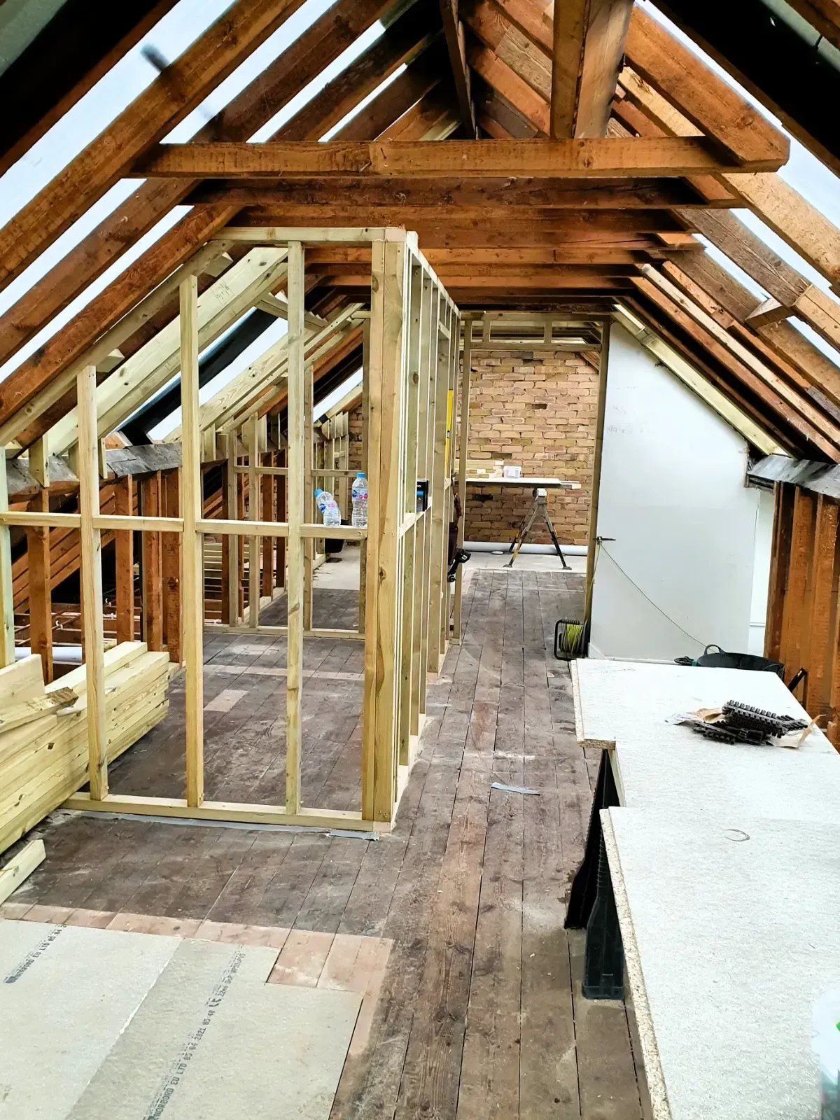 Attic renovation with exposed wooden framing, brick wall, and unfinished floor under a sloped roof
