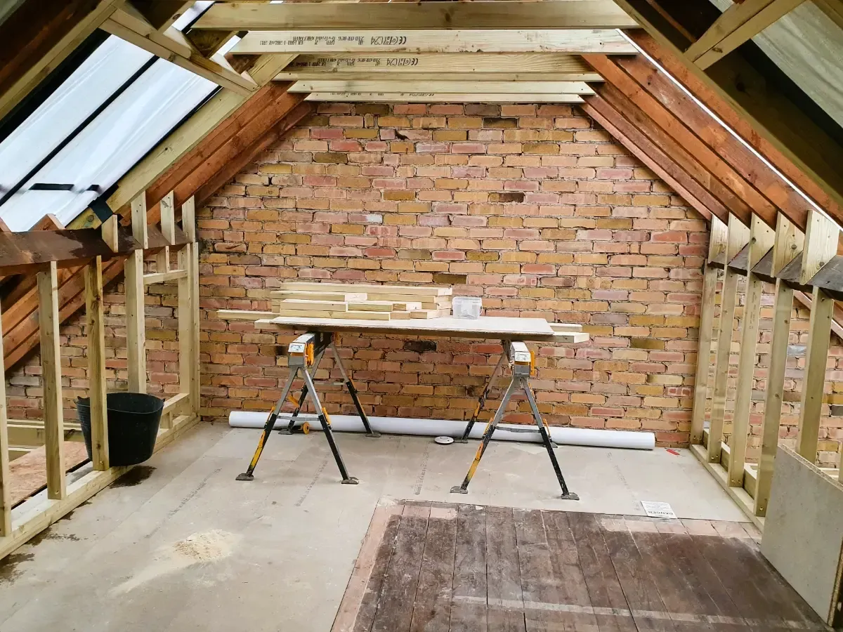 Unfinished loft with exposed brick wall, roof beams, workbench, and ladder under construction.