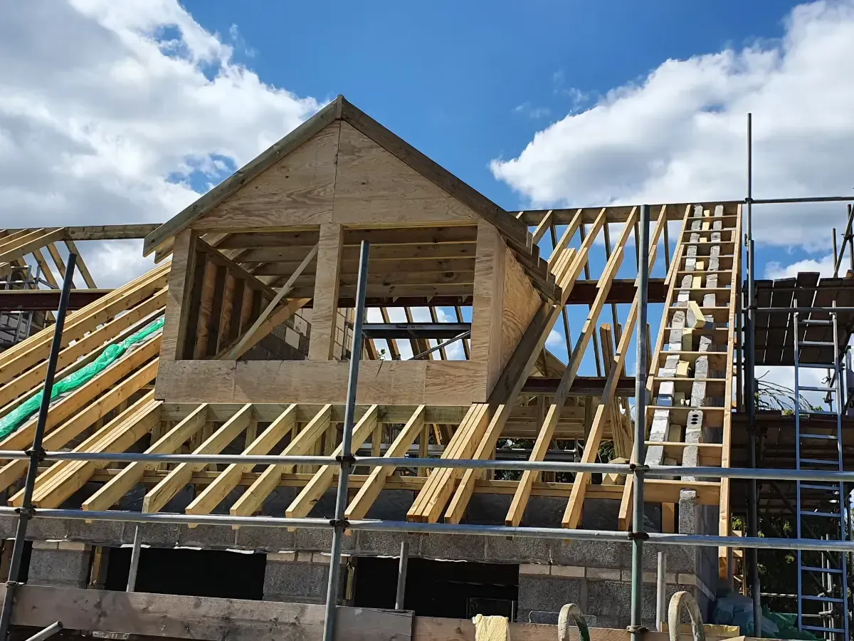 Wood-framed house under construction with exposed roof trusses and scaffolding under a partly cloudy sky