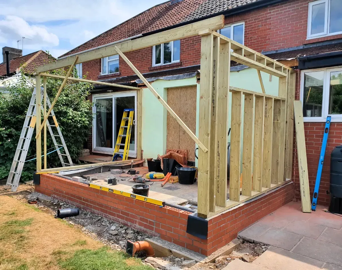 Backyard home extension under construction, with timber framing and tools beside a brick house