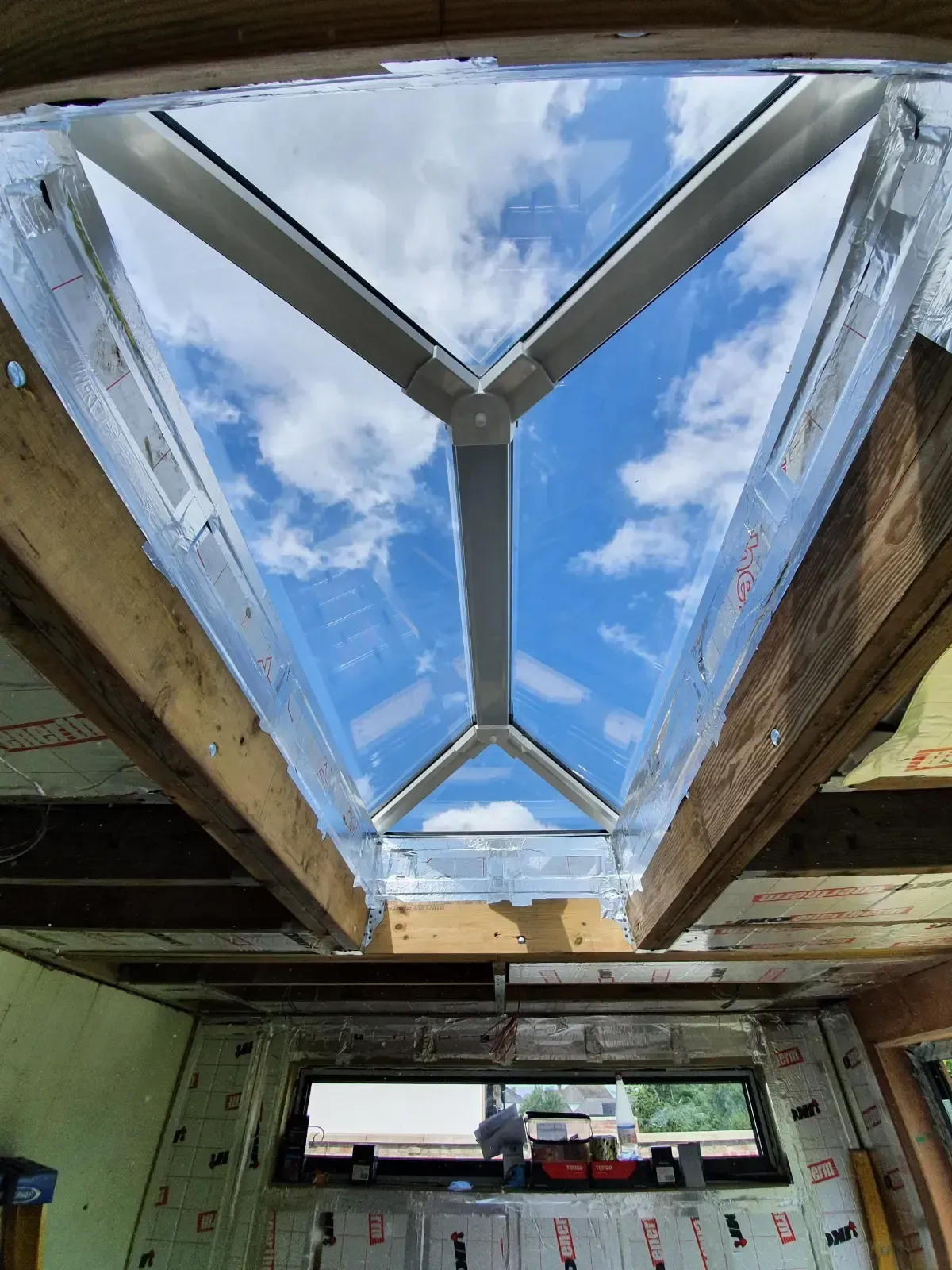 View through a skylight opening with blue sky and clouds, framed by exposed wooden beams inside a building