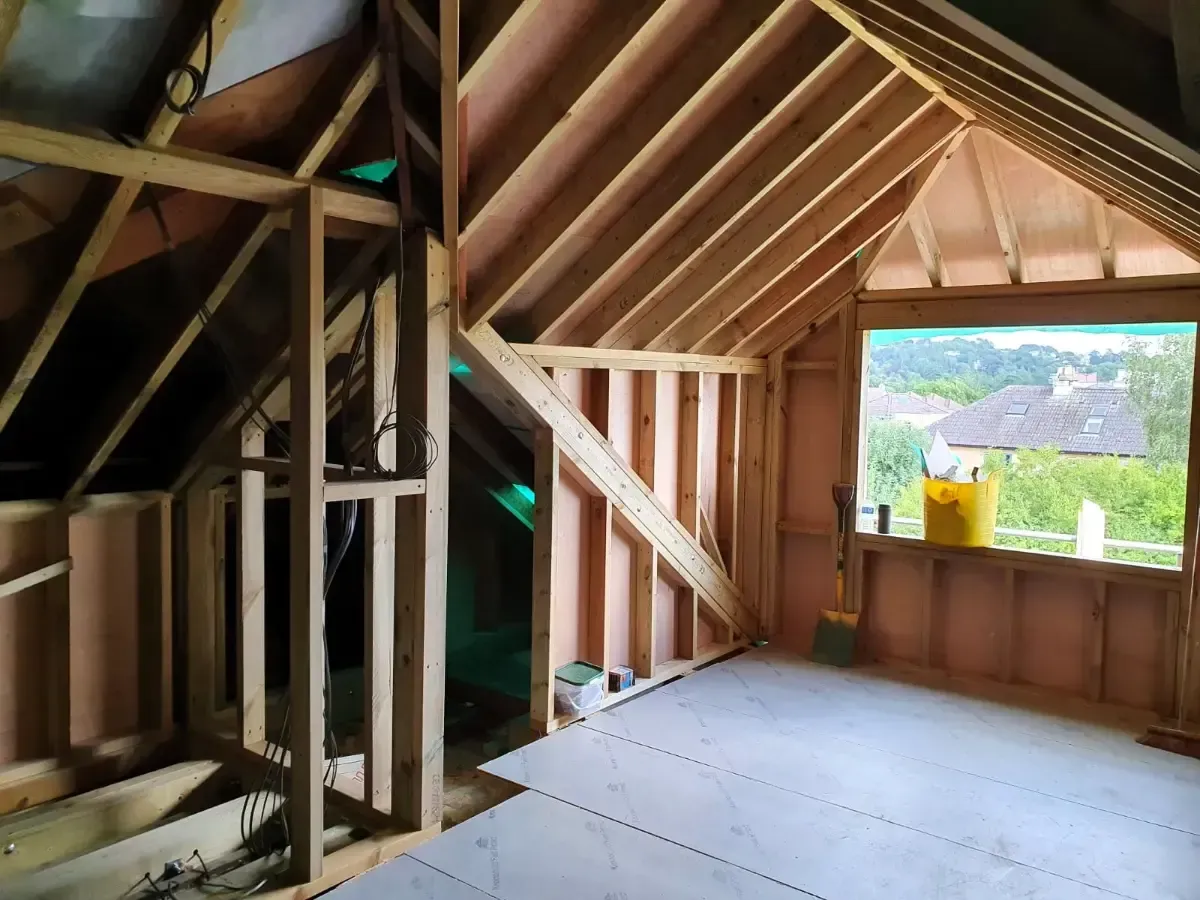 Attic interior under construction with exposed wooden framing and a window overlooking a grassy landscape