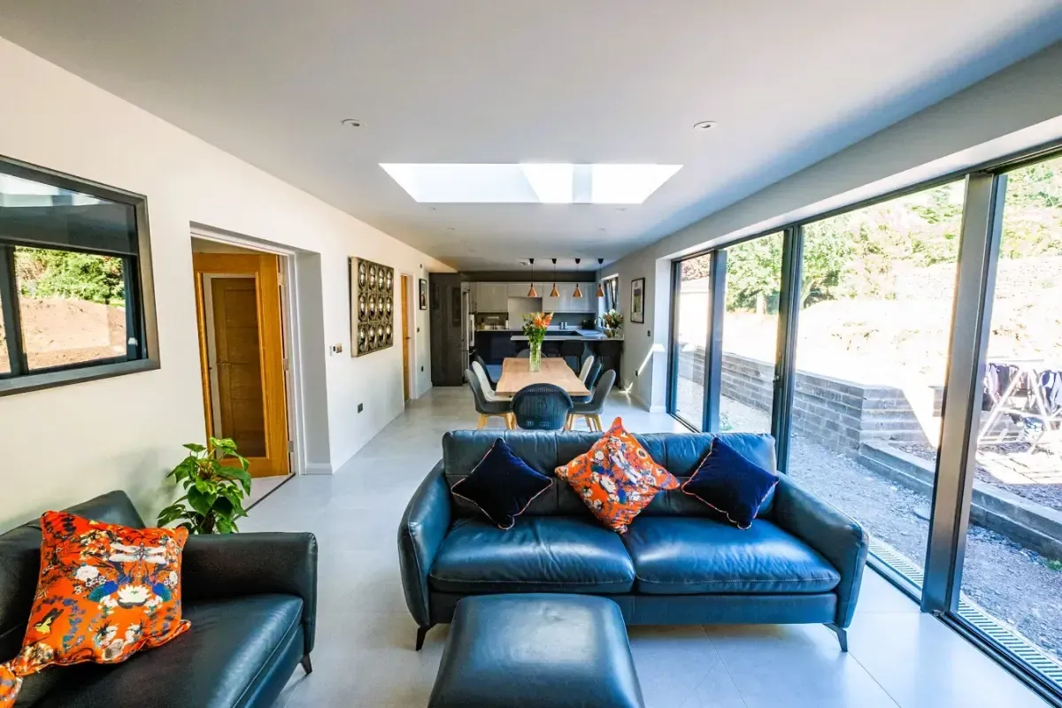Bright living room with black leather sofa, orange accents, skylight, and large glass doors opening to greenery