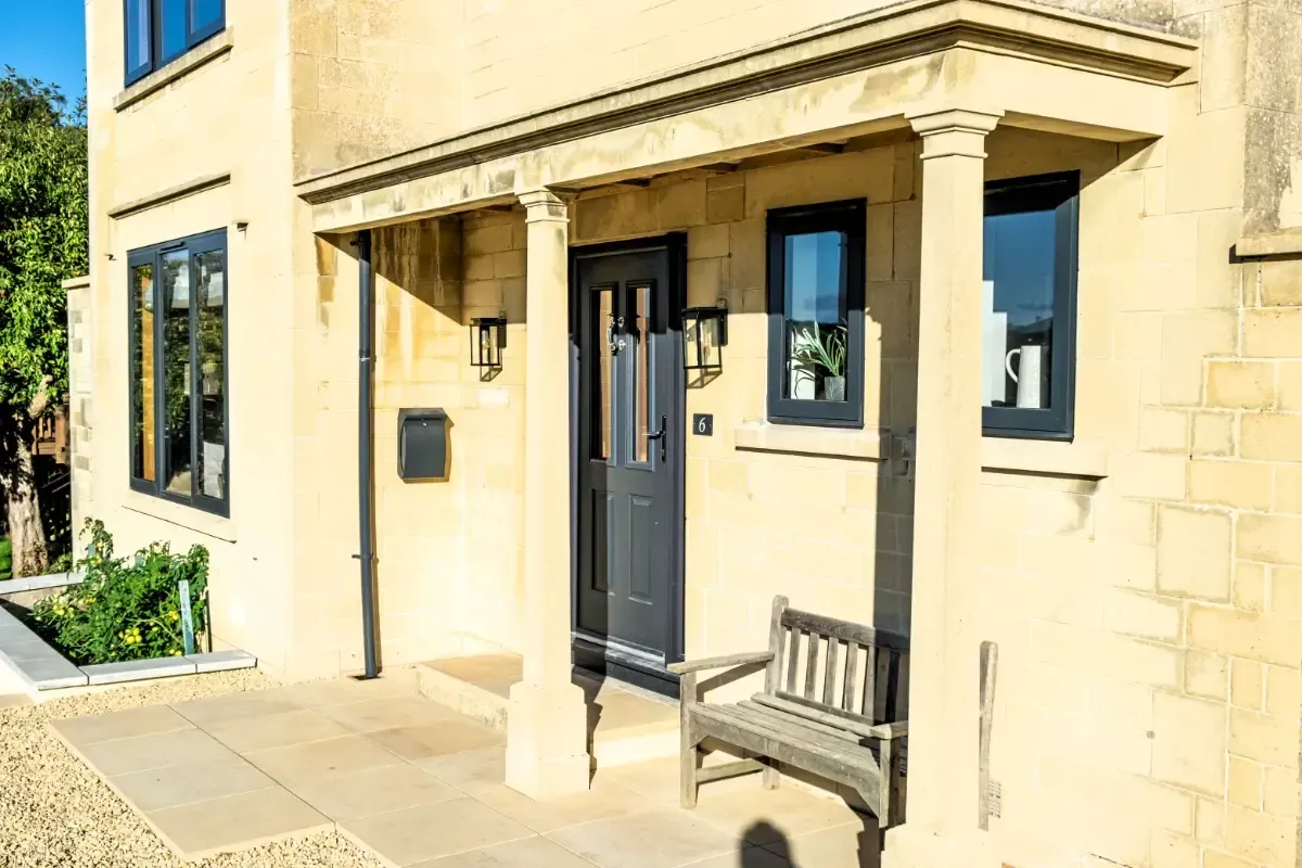 Sunlit stone house entrance with a black door, porch columns, and a wooden bench