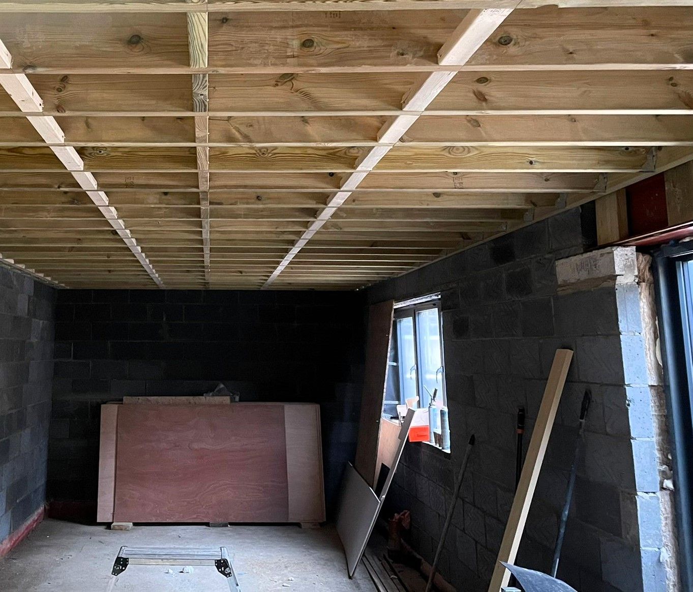 Interior under construction with exposed wooden ceiling joists, cinderblock walls, and boards leaning by a window.