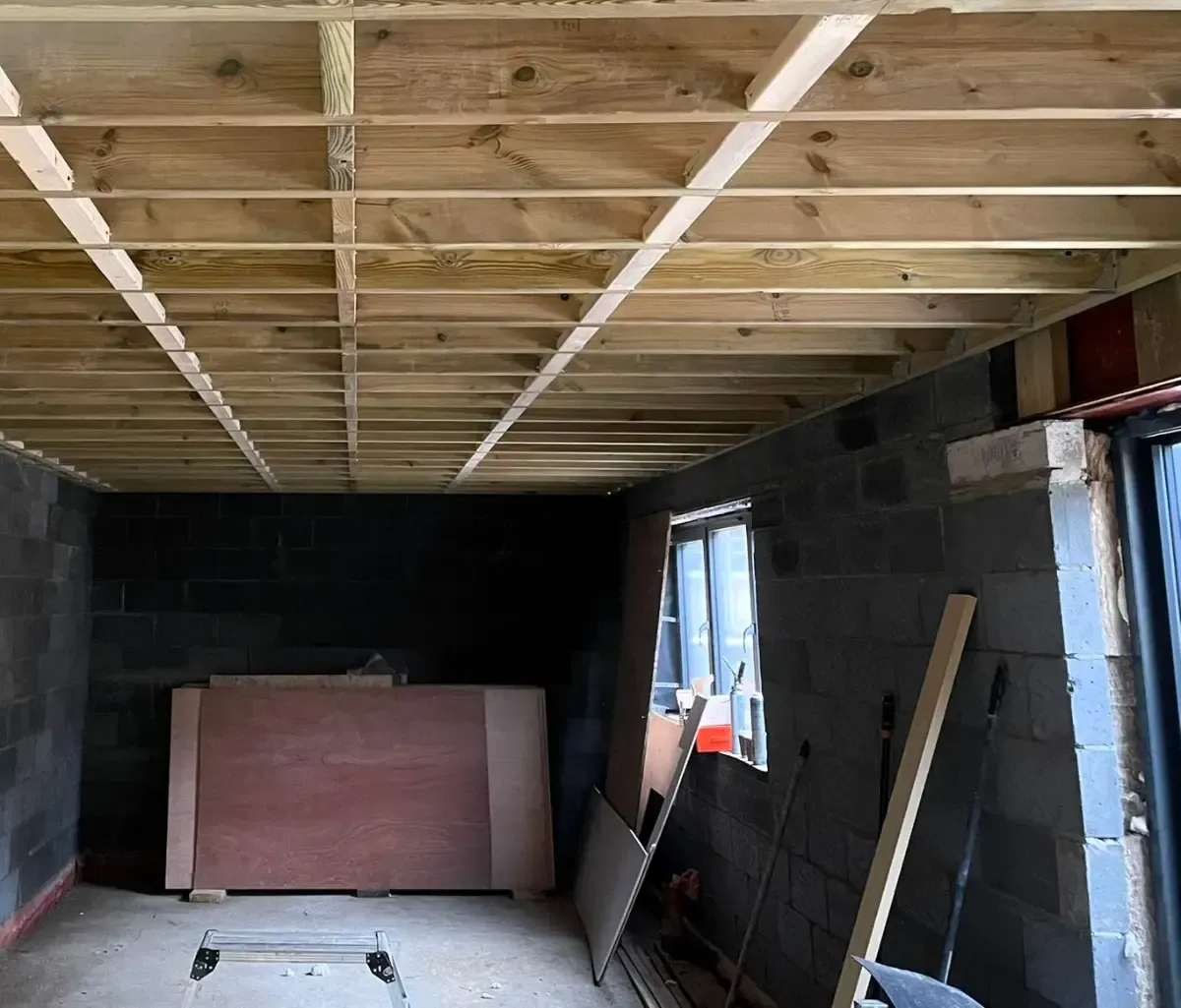 Unfinished room with exposed ceiling joists, cinderblock walls, and construction materials leaning against the wall