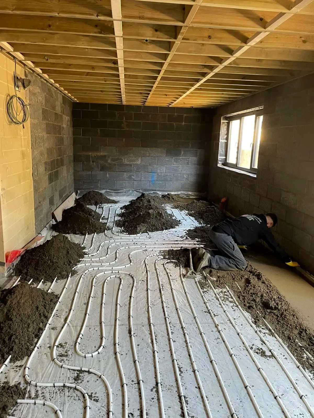 Interior room under construction with exposed ceiling beams, cinderblock walls, and wet floor heating pipes.