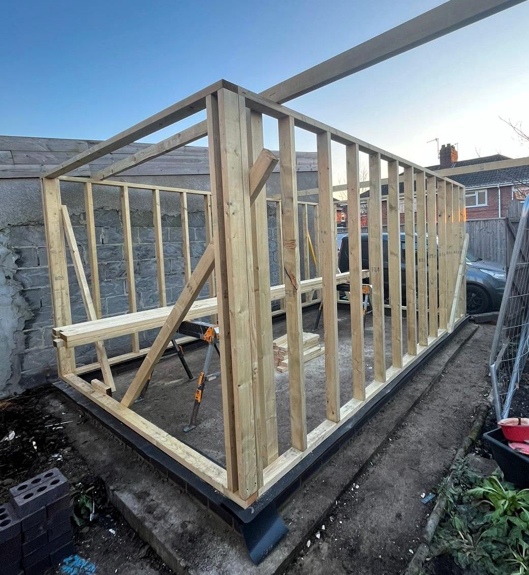Wooden shed frame under construction on a concrete base, with roof beams and vertical studs visible.