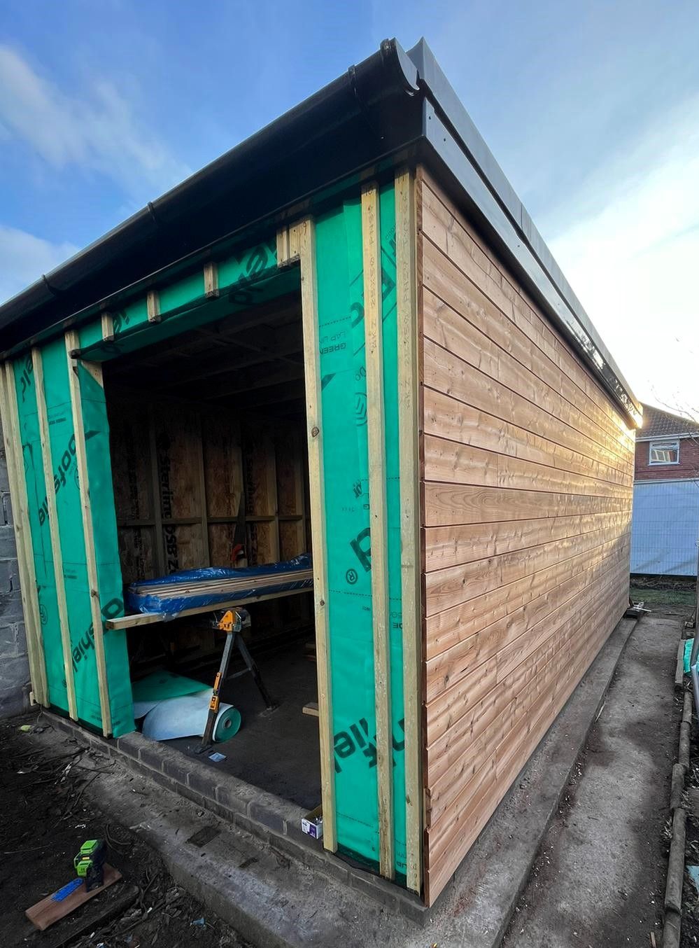 Green wooden shed with open doorway, revealing a small interior and workbench under a cloudy sky