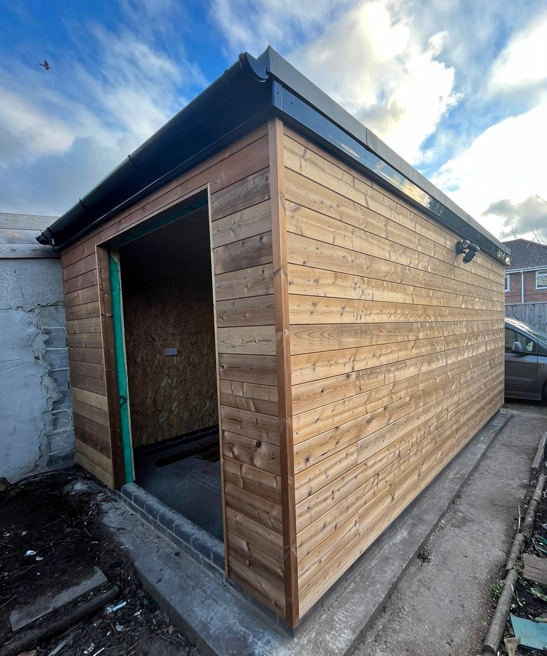 Wooden shed under construction with open doorway and black roof beside a concrete path