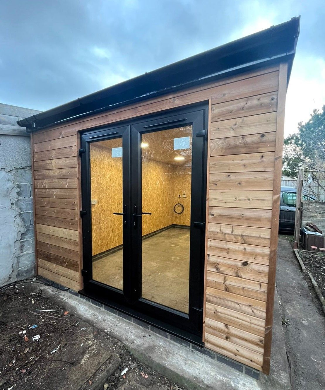 Small wooden garden office with black-framed glass double doors and warm interior lights