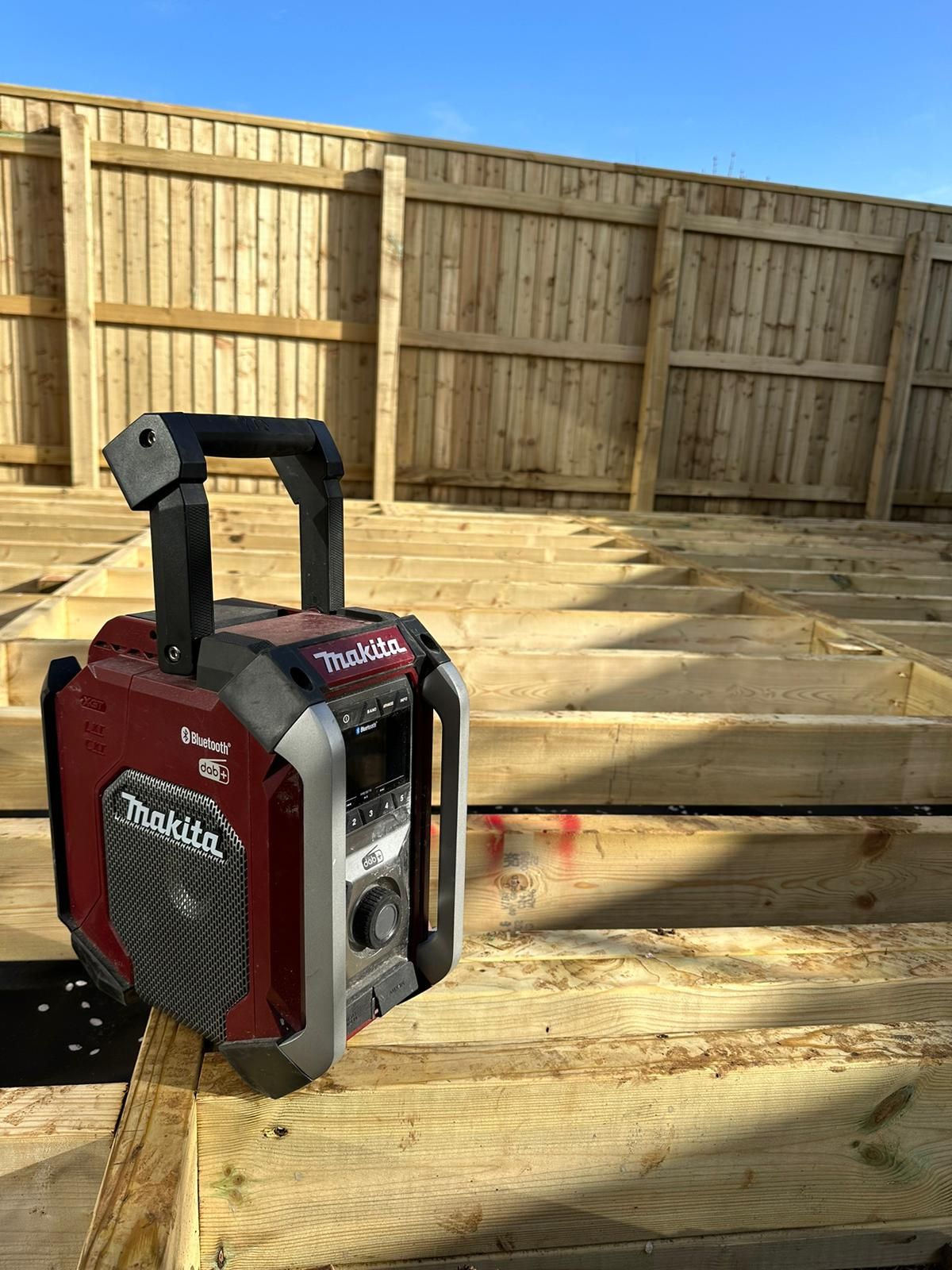 Red and black portable speaker on wooden decking beside a fenced backyard under construction
