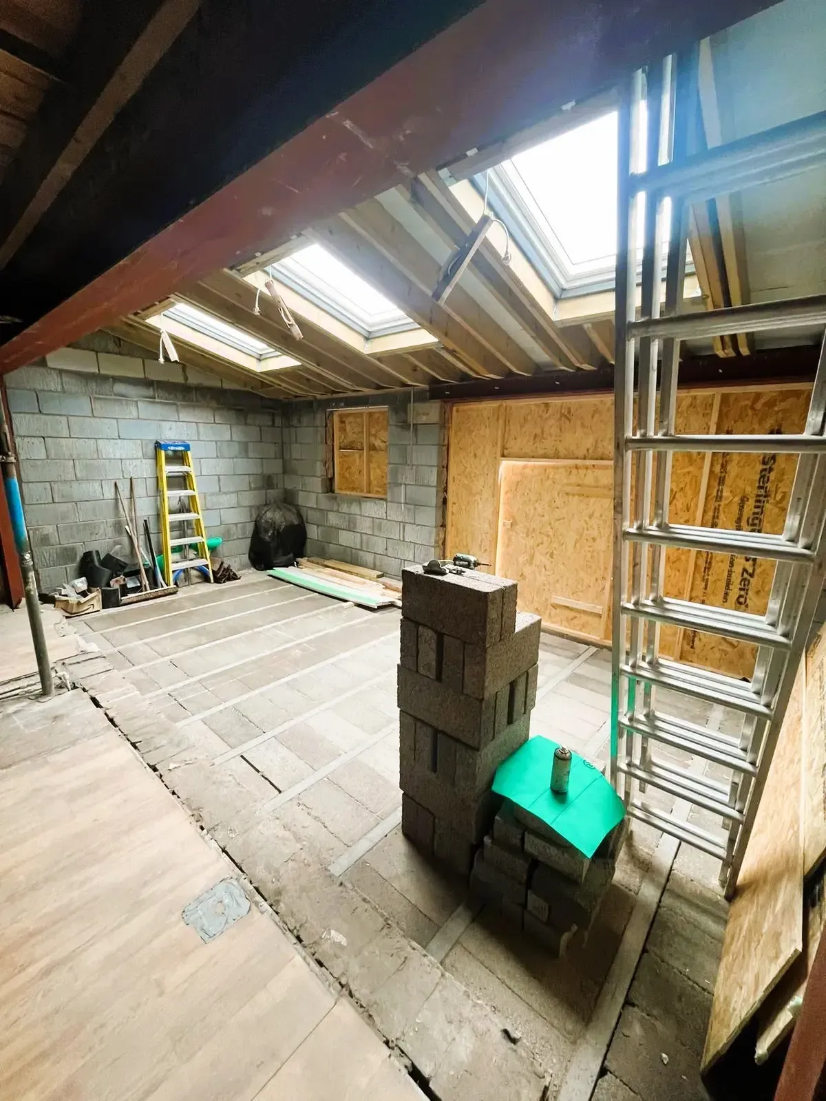 Interior of a partially renovated attic with exposed beams, plywood walls, ladders, and construction materials.