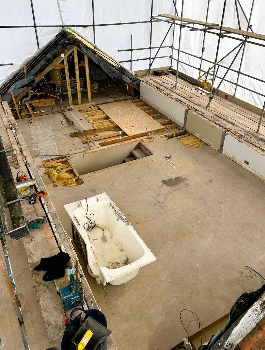 Scaffolding around a roof under repair, with exposed wood, tarps, and a white bathtub on the floor