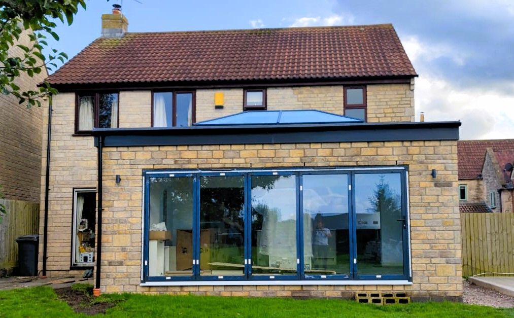 Two-story stone house with red tiled roof and large blue-framed glass extension in front