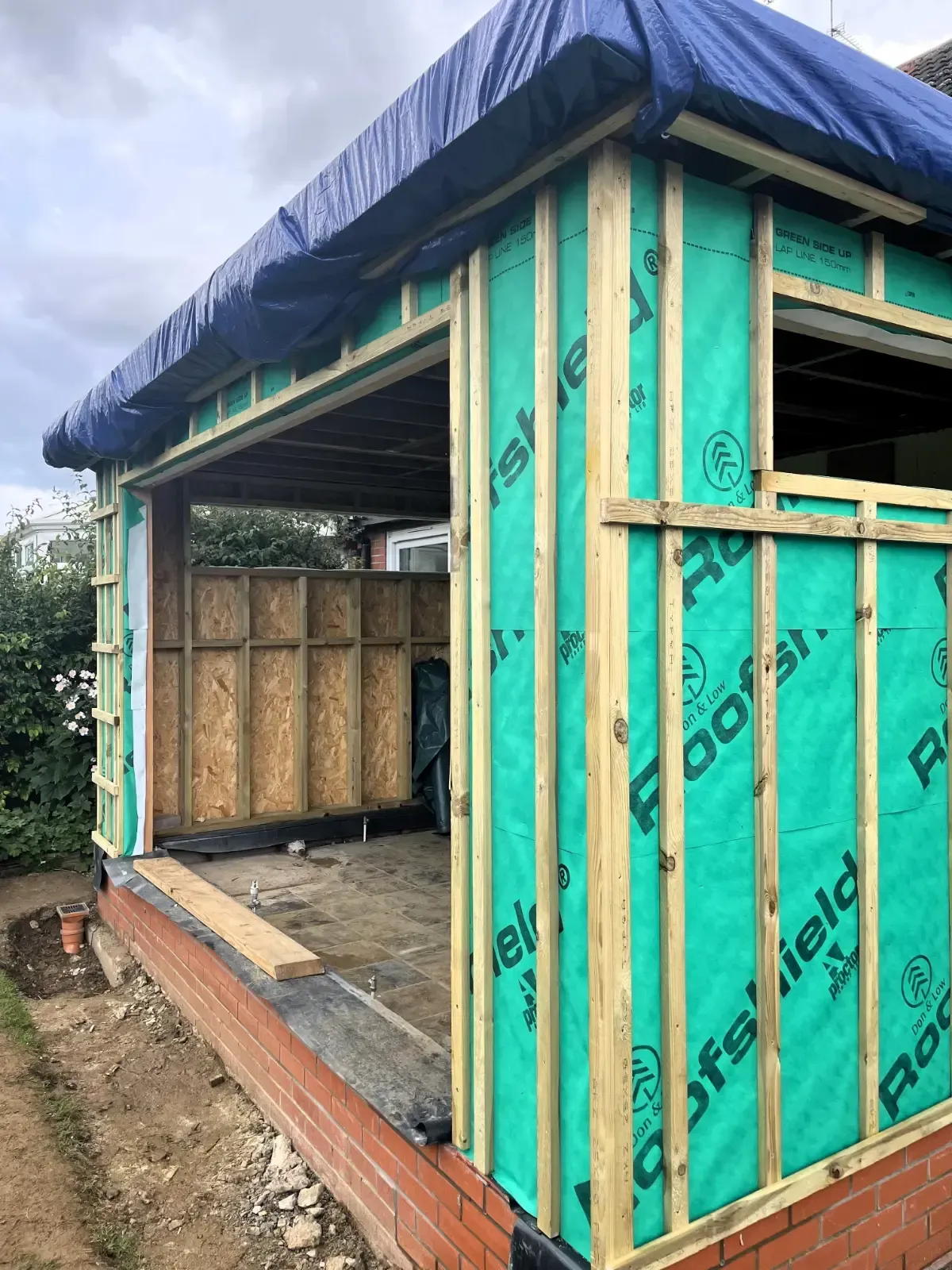 Small unfinished wooden shed with green tarp siding and exposed framing outdoors