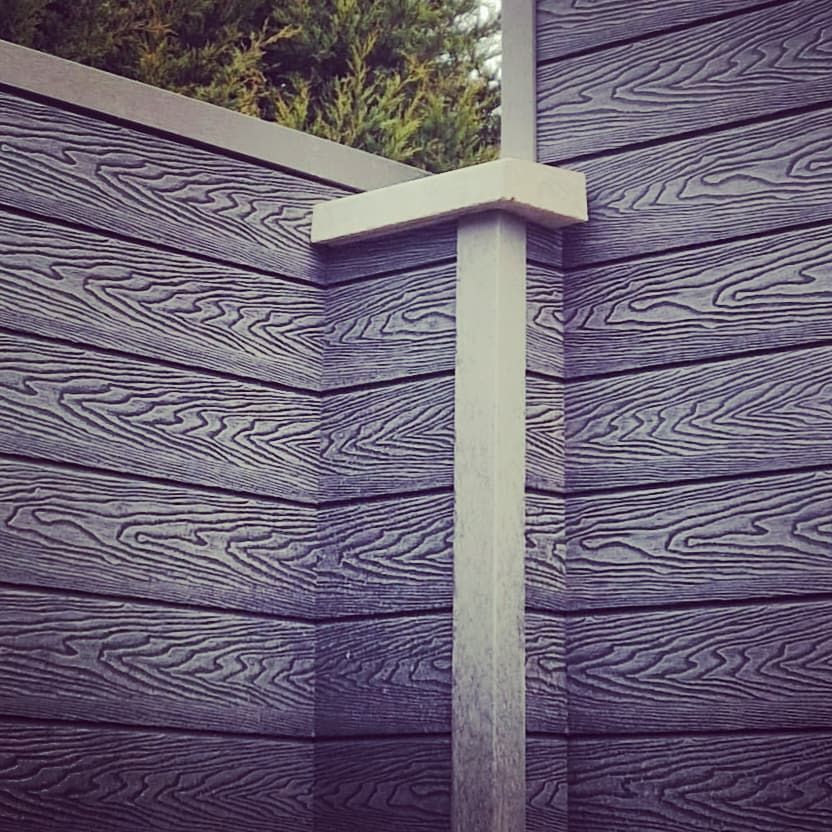 Gray wood-grain fence with a white vertical post and cap against greenery in the background