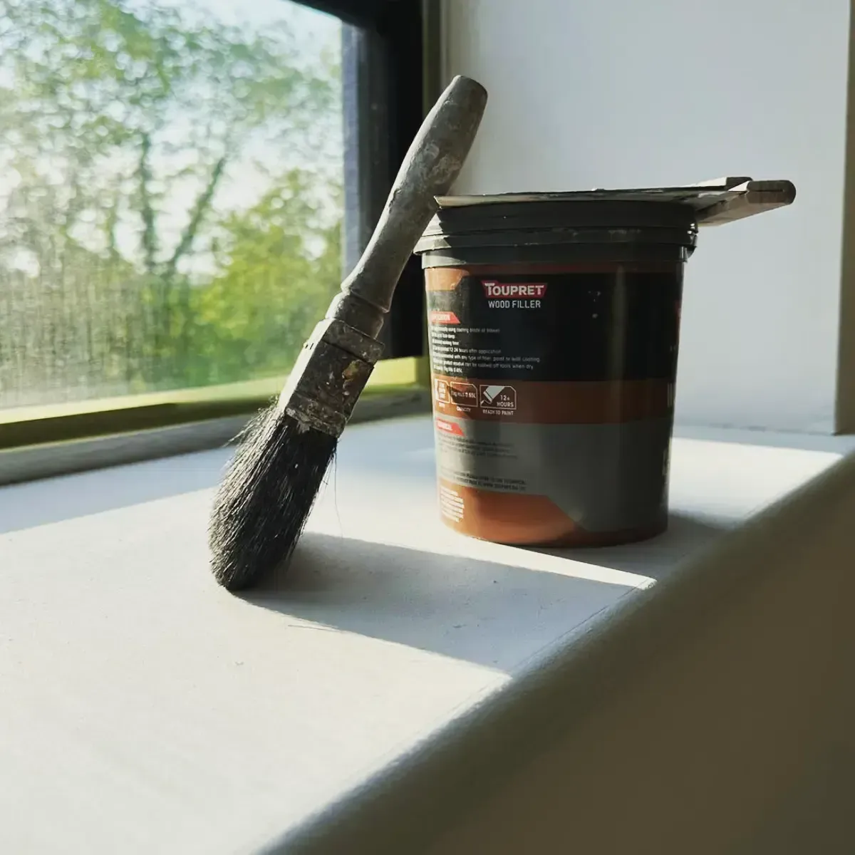 Paint brush leaning on a black and orange paint can beside a sunlit window