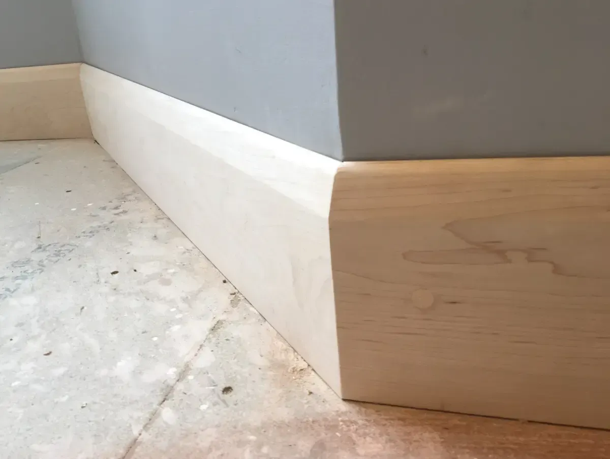 Baseboard trim at a corner where white and light wood meet a gray wall on a tiled floor