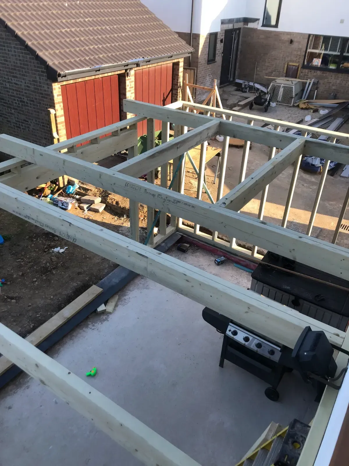 Roof frame under construction beside a house, with gray beams and scattered tools in a backyard.