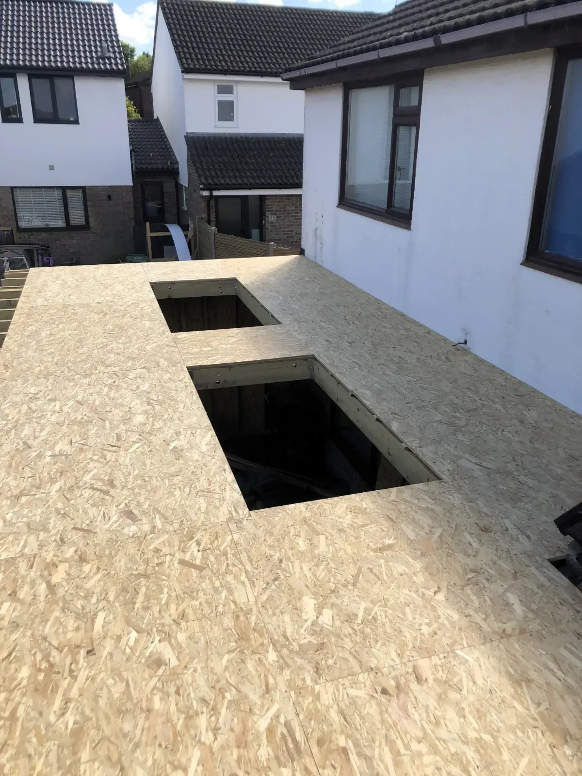 Roof under construction with OSB sheathing and two rectangular openings in a residential courtyard.