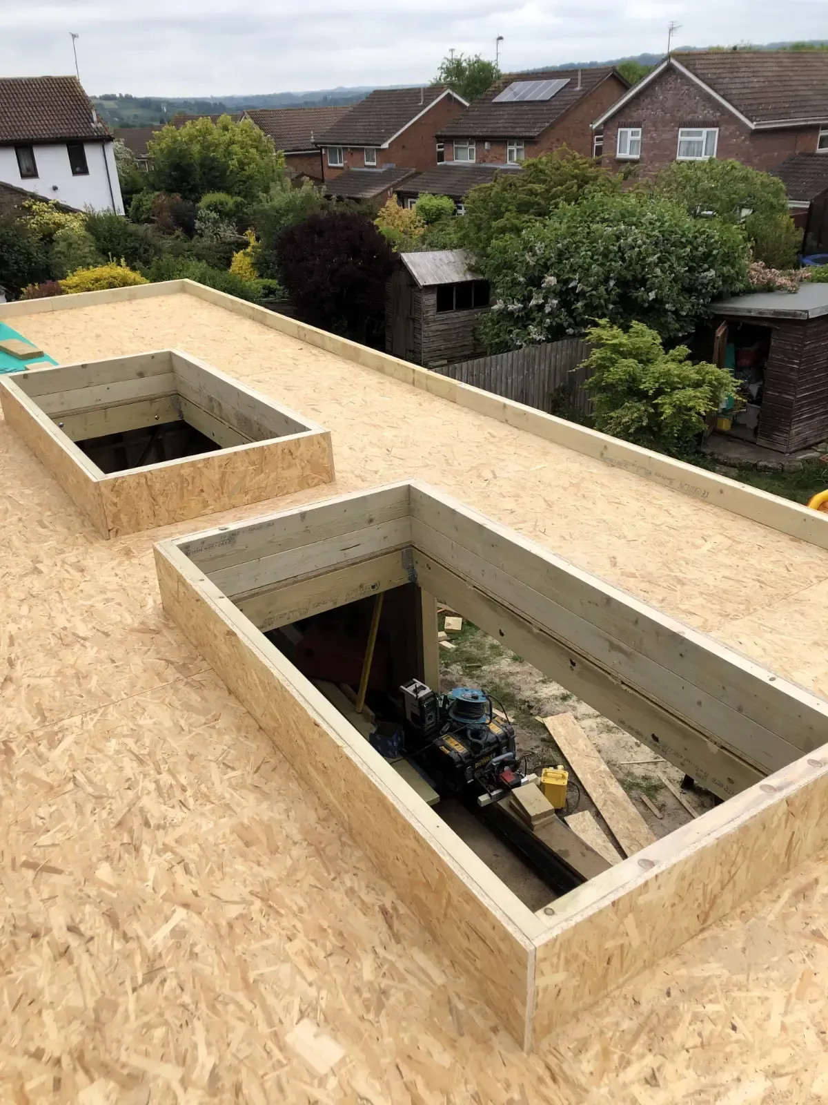 Rooftop under construction with two open skylight openings and suburban houses in the background