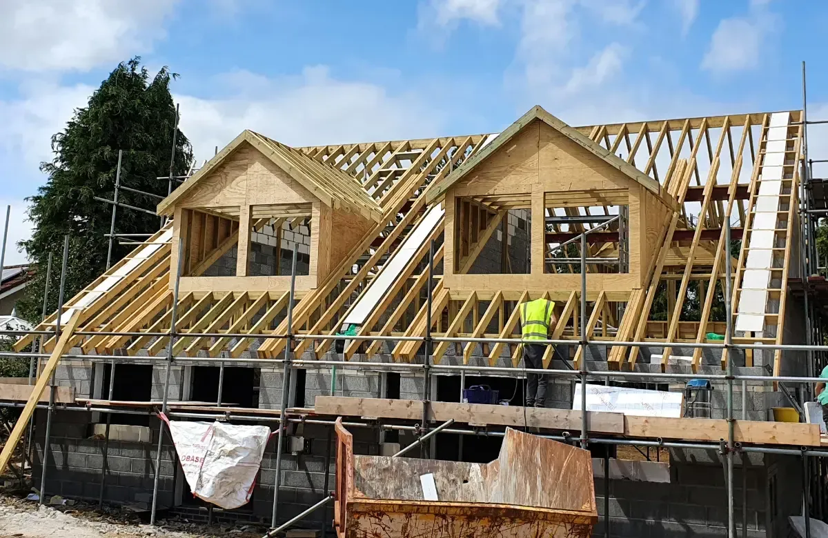 House under construction with exposed wooden roof framing and workers on scaffolding