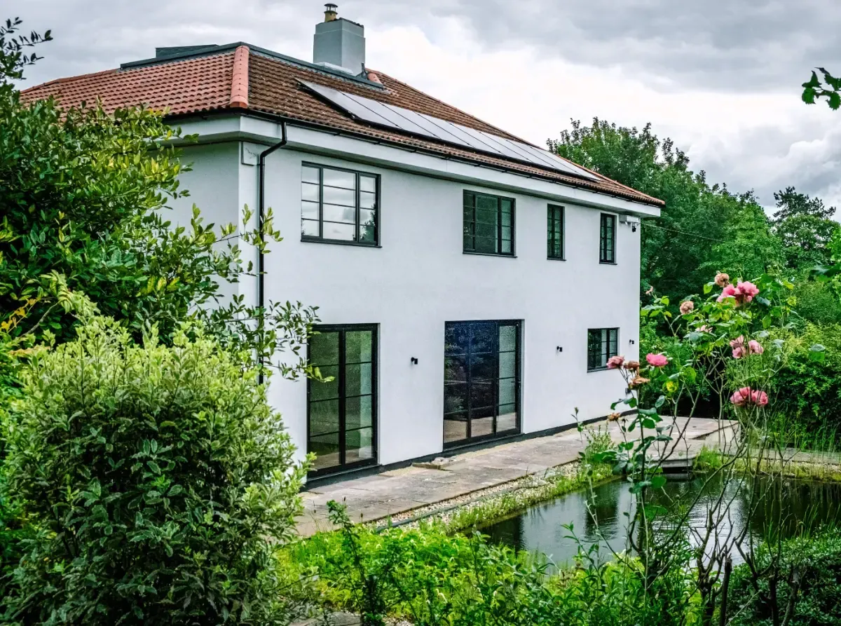 White two-story house with red roof tiles, green window shutters, and lush garden by a pond.