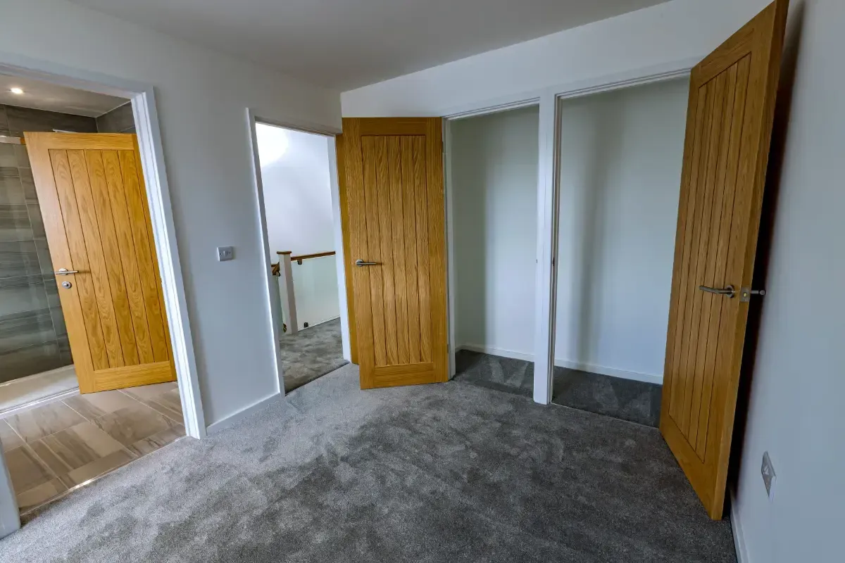 Empty carpeted bedroom with white walls, open wooden doors, and a small adjoining bathroom visible.