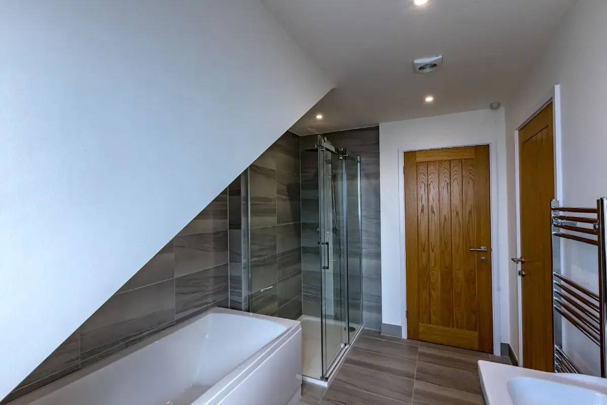 Small bathroom with a glass shower, white bathtub, wood door, and sloped ceiling.