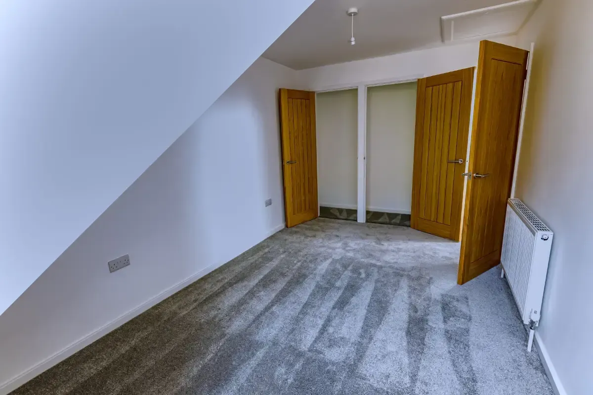 Empty attic bedroom with sloped ceiling, carpeted floor, open wardrobe, and radiator.