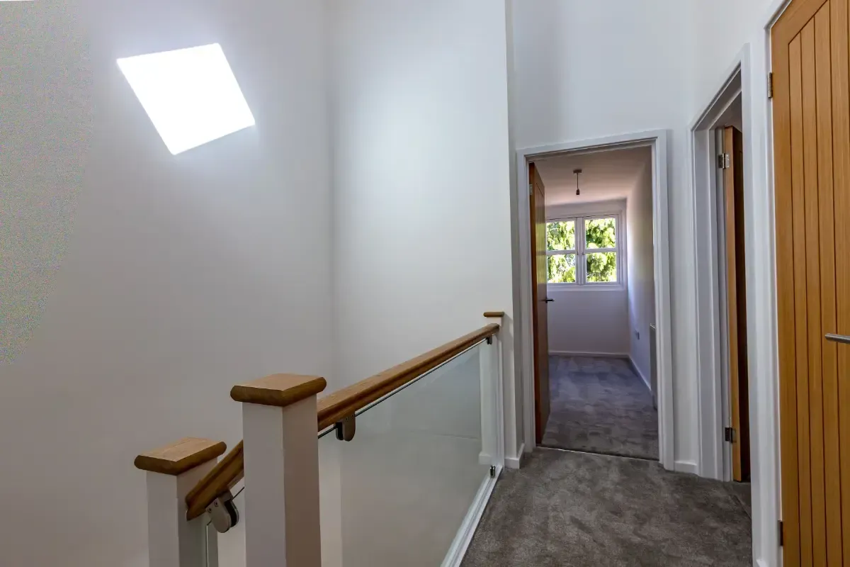 Bright upstairs hallway with skylight, carpeted landing, wooden railings, and a doorway to a room with a window.