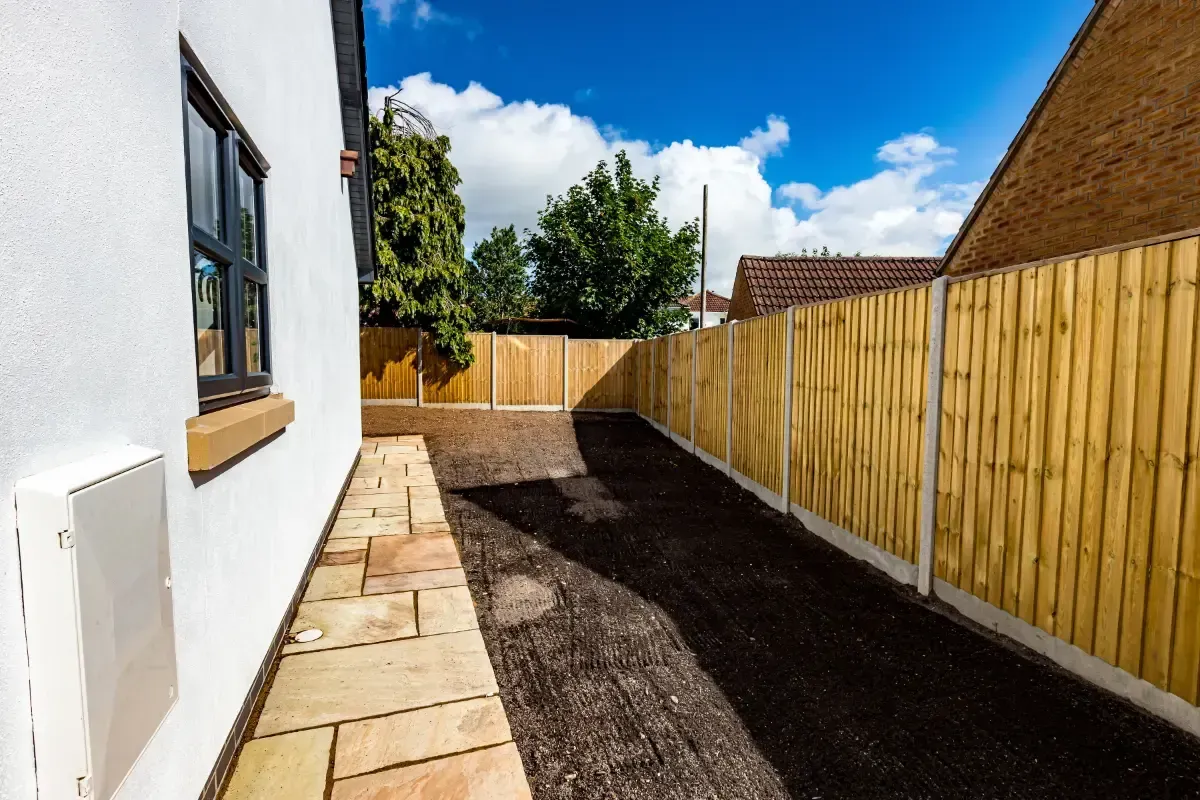 Narrow side yard with fresh soil, wooden fence, and white house wall under a bright blue sky