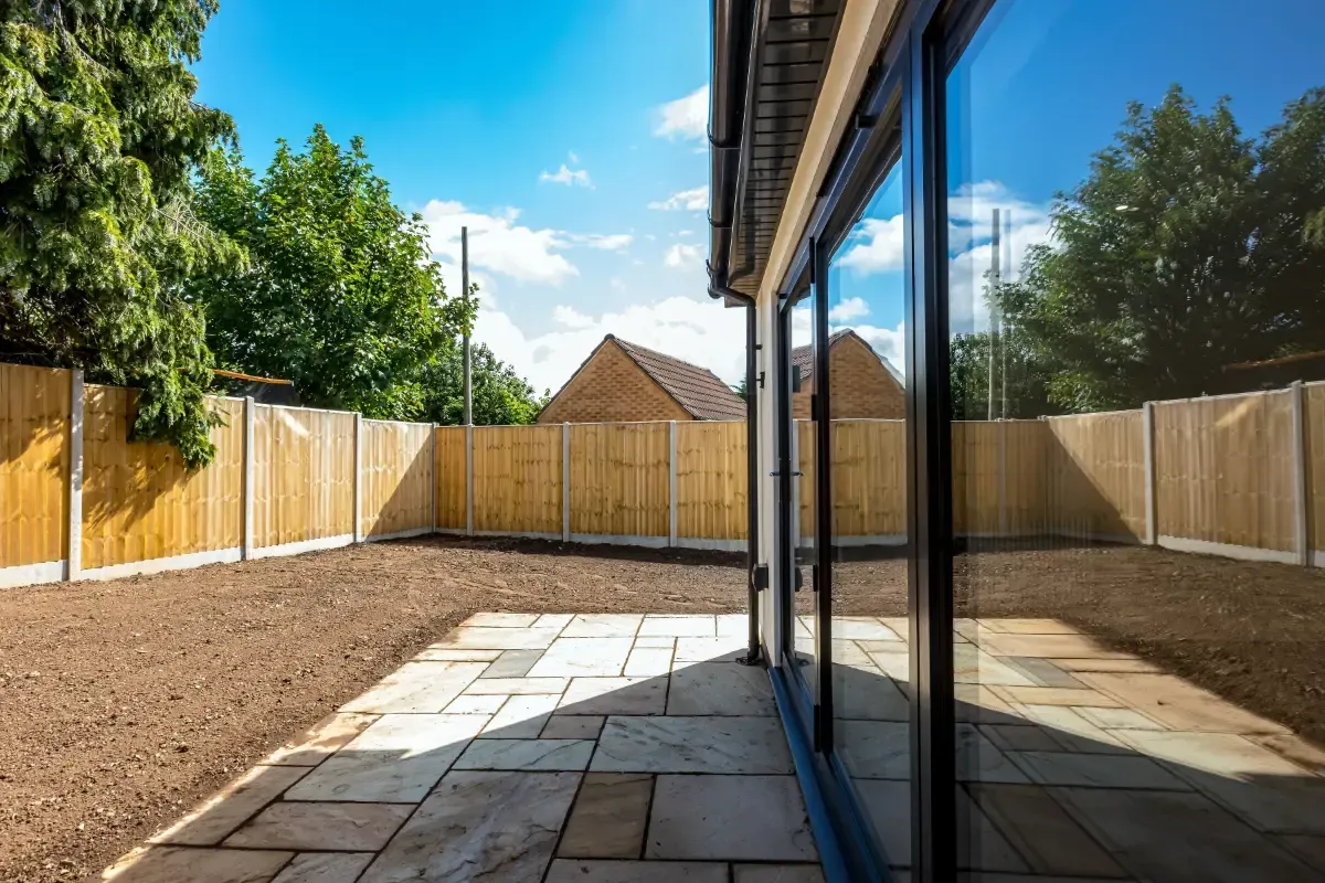 Backyard patio with stone paving, gravel, wooden fence, and glass house wall under a blue sky
