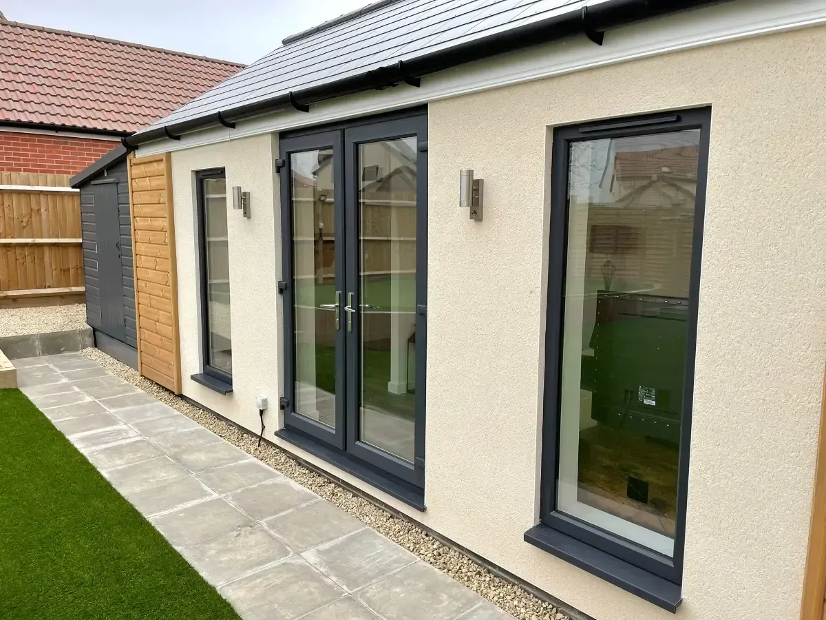 Modern cream house exterior with dark-framed patio doors and windows, viewed along a paved side path