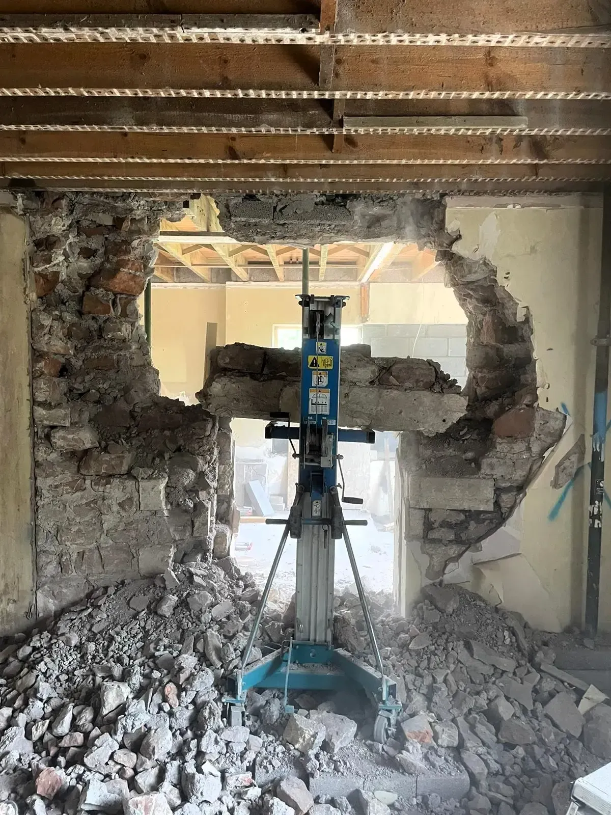 Demolition site with a jackhammer drilling a large hole in a crumbling wall, debris piled on the floor.