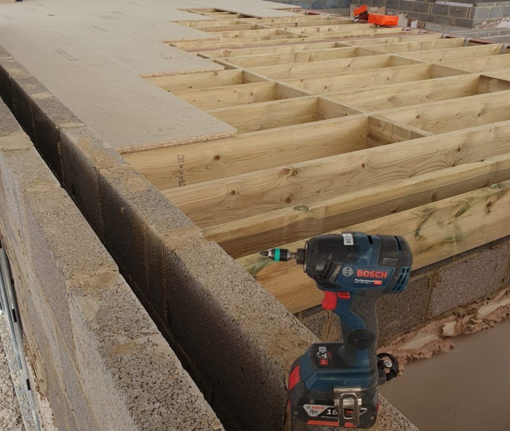 Power drill on a wooden deck frame beside a concrete edge, with orange tools in the background.