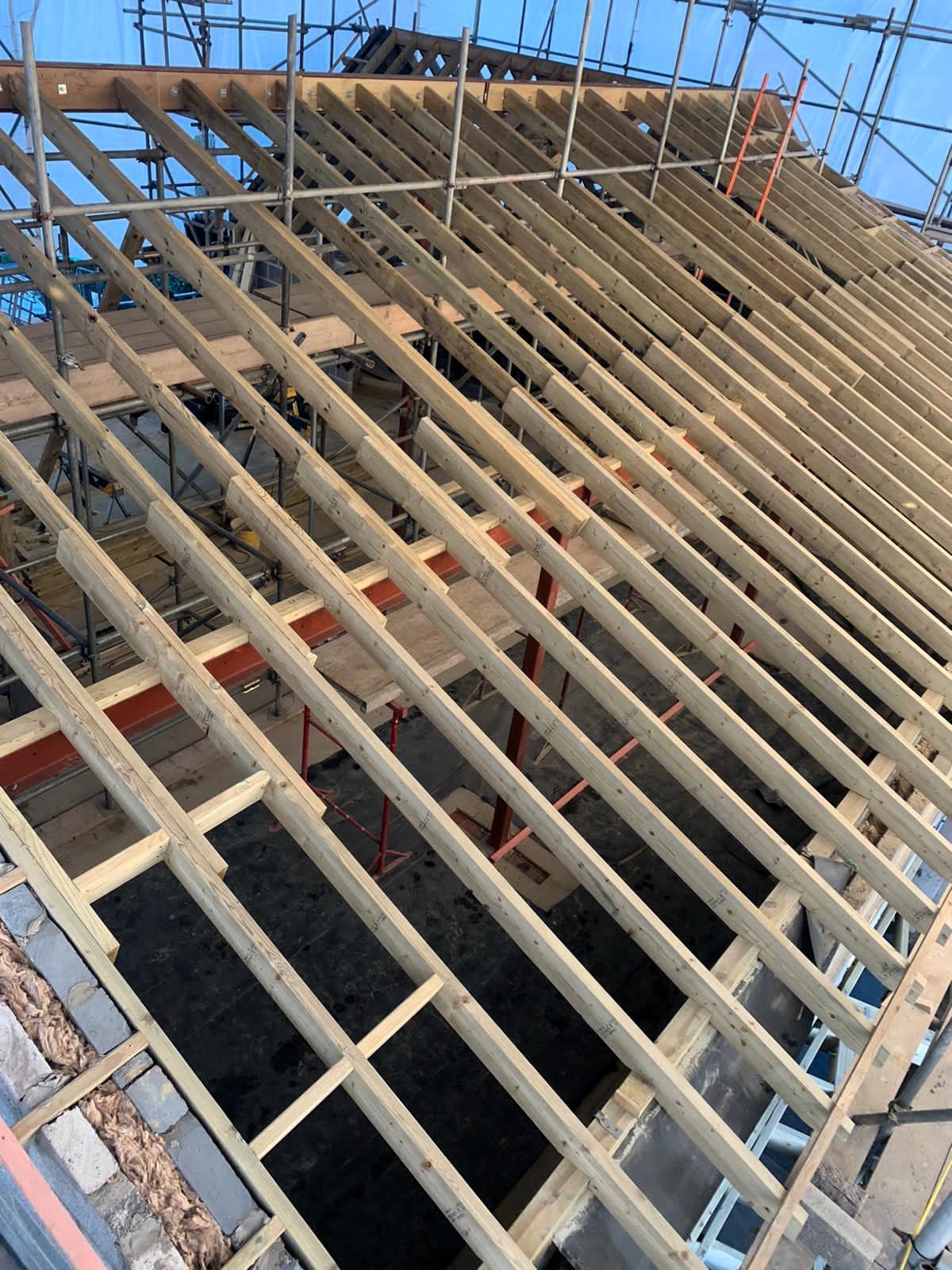 Wooden roof framing over a building under construction, with blue sky visible through the beams