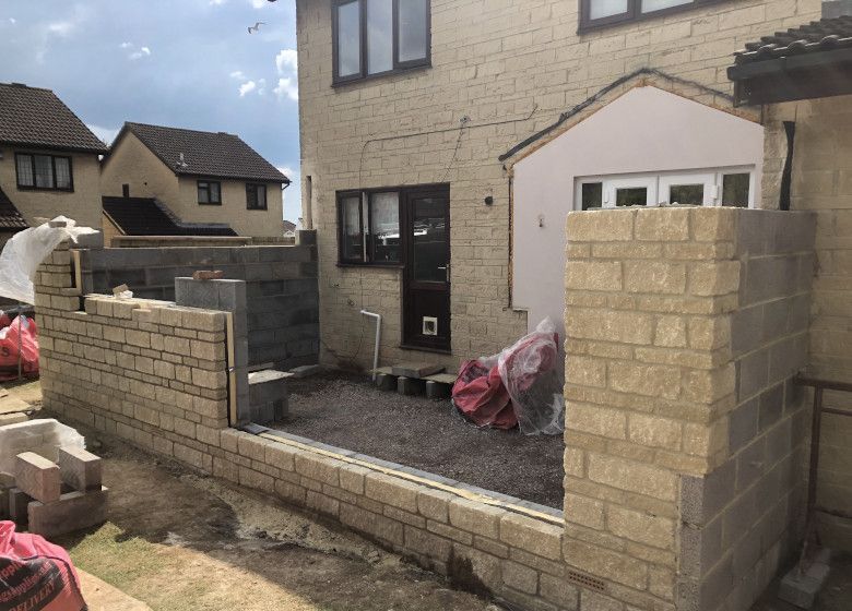 Brick front wall under construction beside a house with workers and building materials in a residential yard