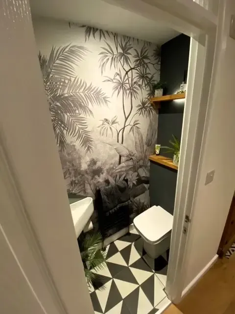 Small bathroom with black-and-white geometric floor, tropical wallpaper, and shelving beside the toilet.