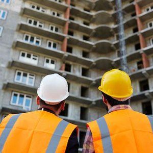 Two construction workers wearing hard hats and orange vests are looking at a building under construction.