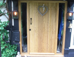 Purpose-made-joinery---Malvern,-Worcester,-Ledbury---Woodman-Joinery---Bespoke-joinery2 A wooden front door with a heart-shaped wreath, flanked by glass panels, black lanterns, and a mailbox on a vine-covered wall.