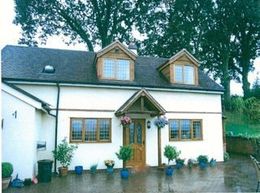 Purpose-made-joinery---Malvern,-Worcester,-Ledbury---Woodman-Joinery---Bespoke-joinery3 A two-story white house with a dark roof, wooden accents, two dormer windows, and a paved front patio with potted plants.