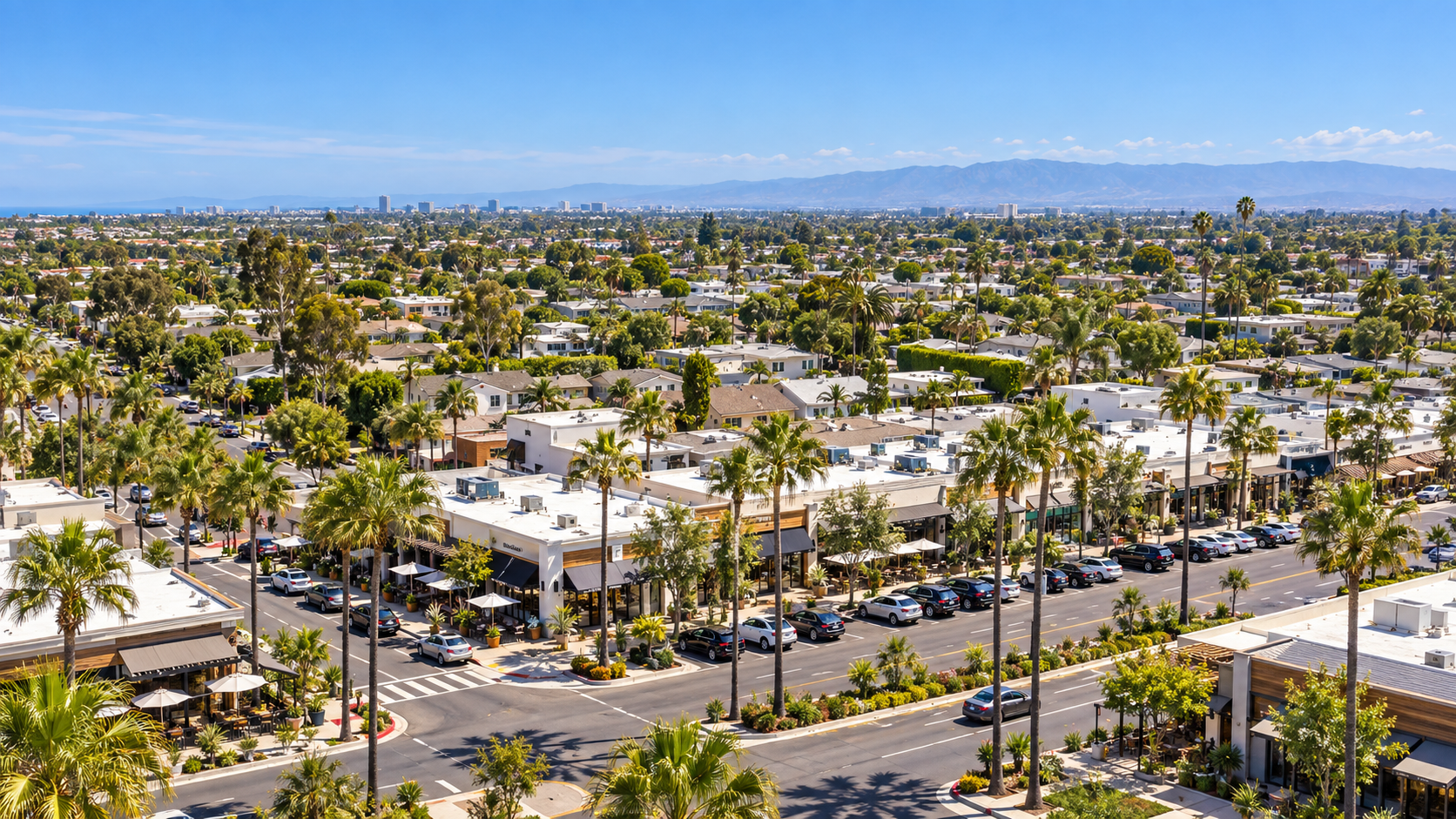 Palm-lined suburban neighborhood with low-rise buildings, parked cars, and distant mountains under a clear blue sky