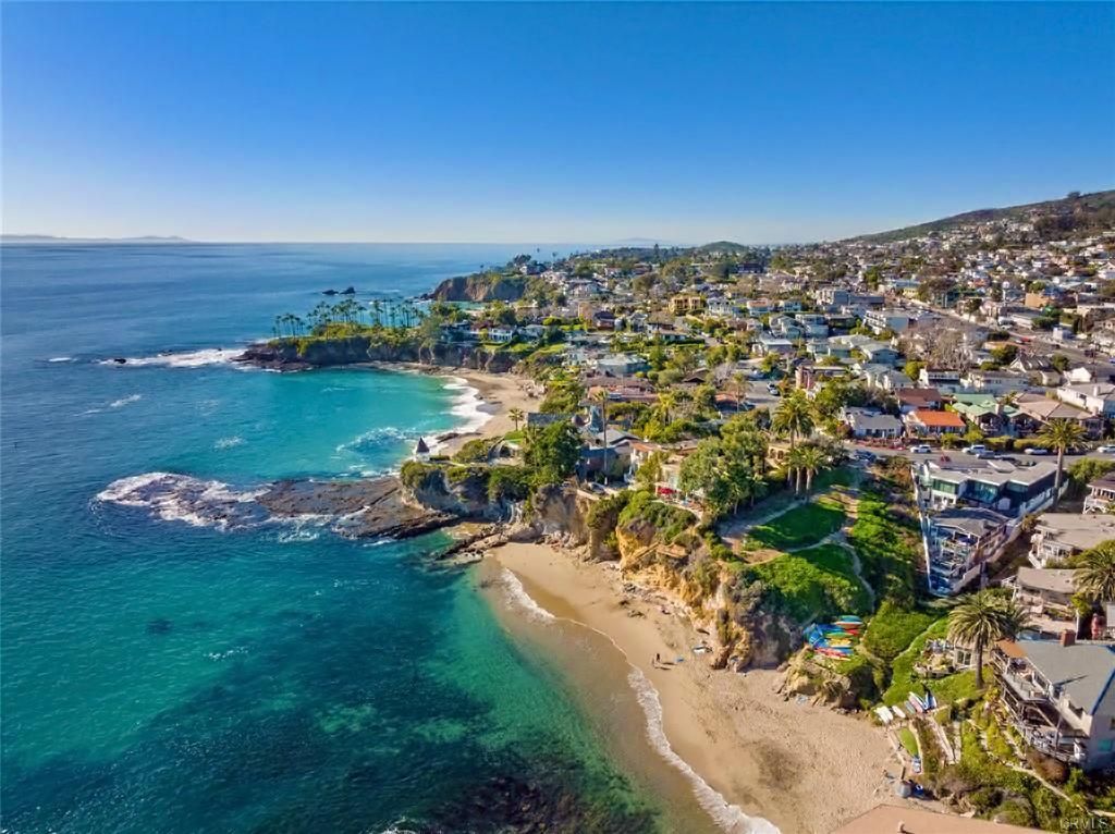 Aerial view of a turquoise beach cove beside a hillside town with houses and cliffs.