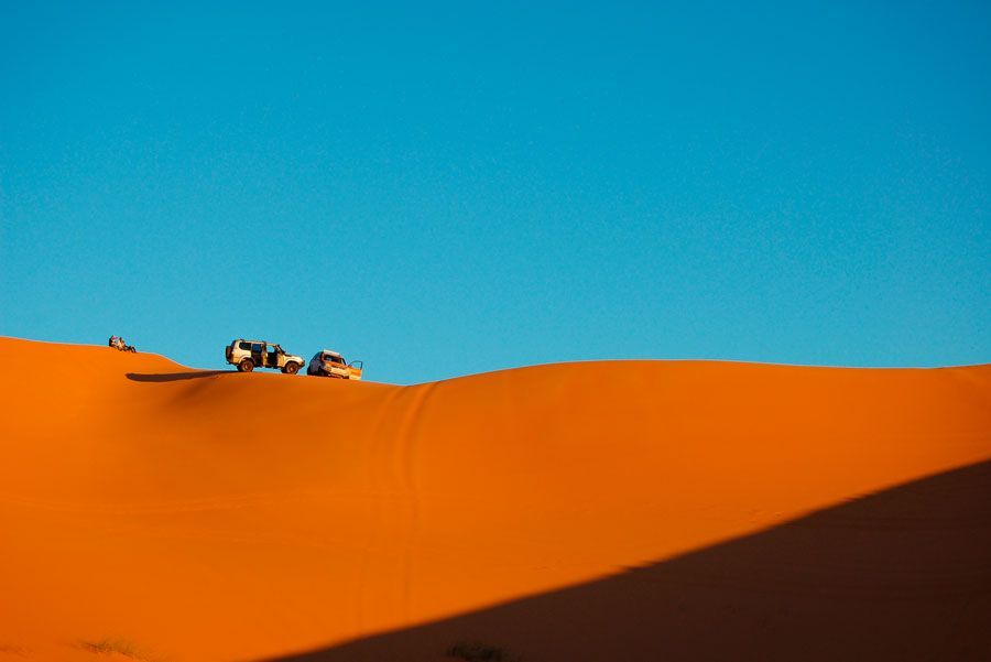 Vehículos atravesando una gran duna de arena naranja bajo un cielo azul claro.