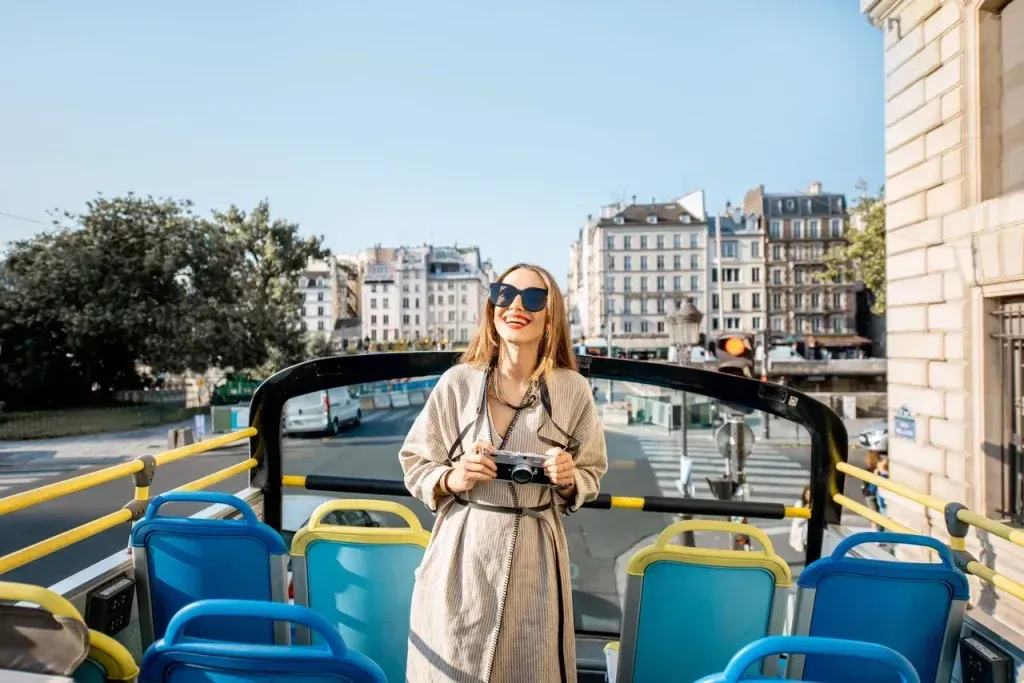 Mujer con gafas de sol y cámara en un autobús descapotable en una ciudad, sonriendo. Edificios y asientos azules al fondo.