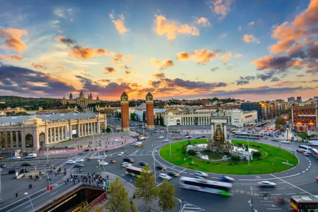 La Plaza de España de Barcelona al atardecer. Los edificios rodean una rotonda concurrida, con tráfico y gente, bajo un cielo colorido.