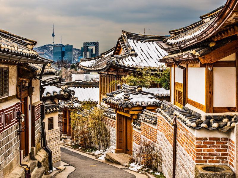 Una calle nevada en el pueblo Bukchon Hanok, Seúl, Corea del Sur, bordeada de casas tradicionales coreanas con techos de tejas y paredes de ladrillo; la Torre N de Seúl a lo lejos.