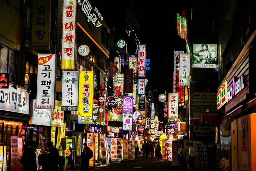 Escena callejera nocturna en Seúl, Corea del Sur, repleta de letreros comerciales iluminados en varios idiomas. La gente camina por la calle.